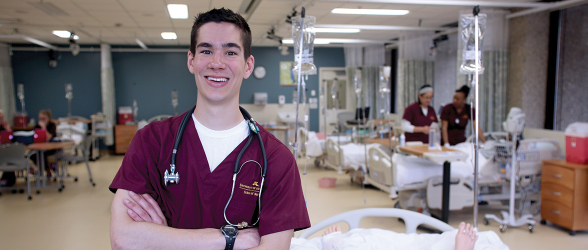 male nursing student in maroon scrubs standing in front of a simulation hospital setting